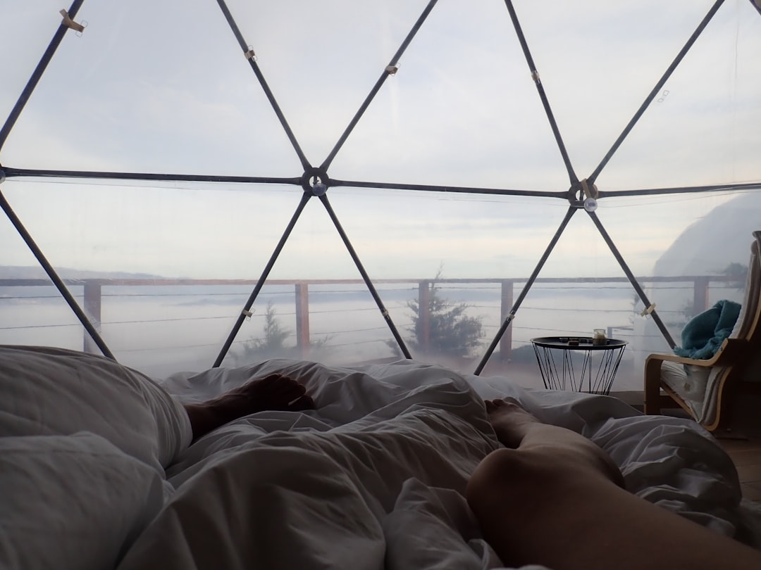 Person lying in bed inside a geodesic dome with large windows, enjoying a Luxus Glamping retreat that overlooks an outdoor deck and a scenic, misty landscape, blending comfort with the beauty of nature.
