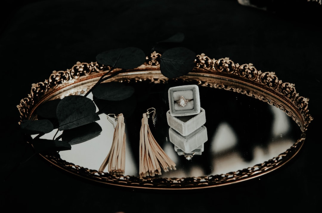 A gold decorative tray with a mirrored surface holds tassel earrings, a leafy branch, and a diamond ring in a gray box—perfect for displaying custom jewelry or showcasing personalized jewelry services.