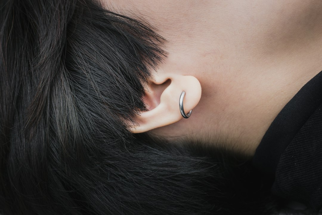 A close-up of a person's ear with short black hair, wearing a small silver hoop earring that adds a touch of high fashion jewelry style.
