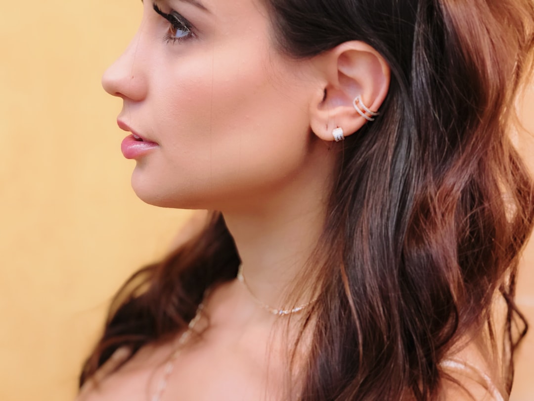 Woman with long brown hair shown in profile, wearing small hoop earrings and a delicate necklace that adds a subtle sparkle, against a plain yellow background.