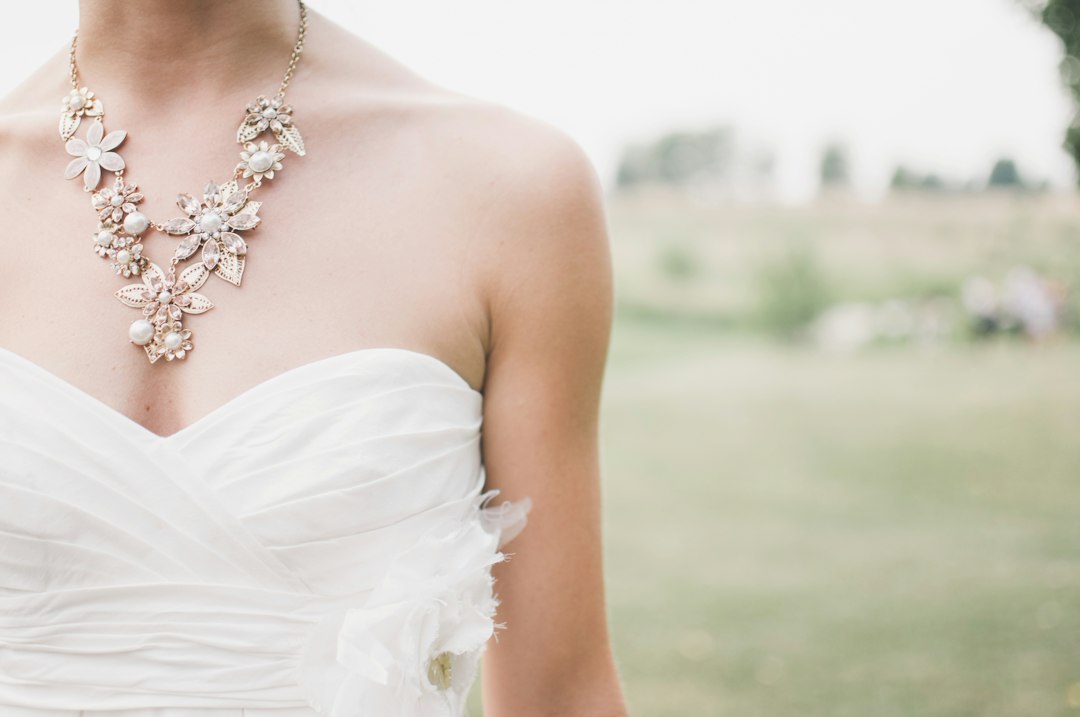 A person in a strapless white dress with a large floral jewelry necklace stands outdoors in a grassy area.