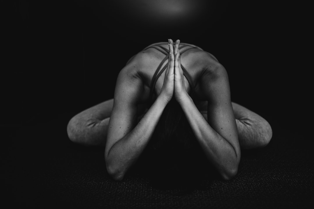 Person in athletic wear kneeling on a mat in a yoga pose, emphasizing the beauty of movement as their arms are bent, palms pressed together, and forehead resting on the floor, against a dark background.