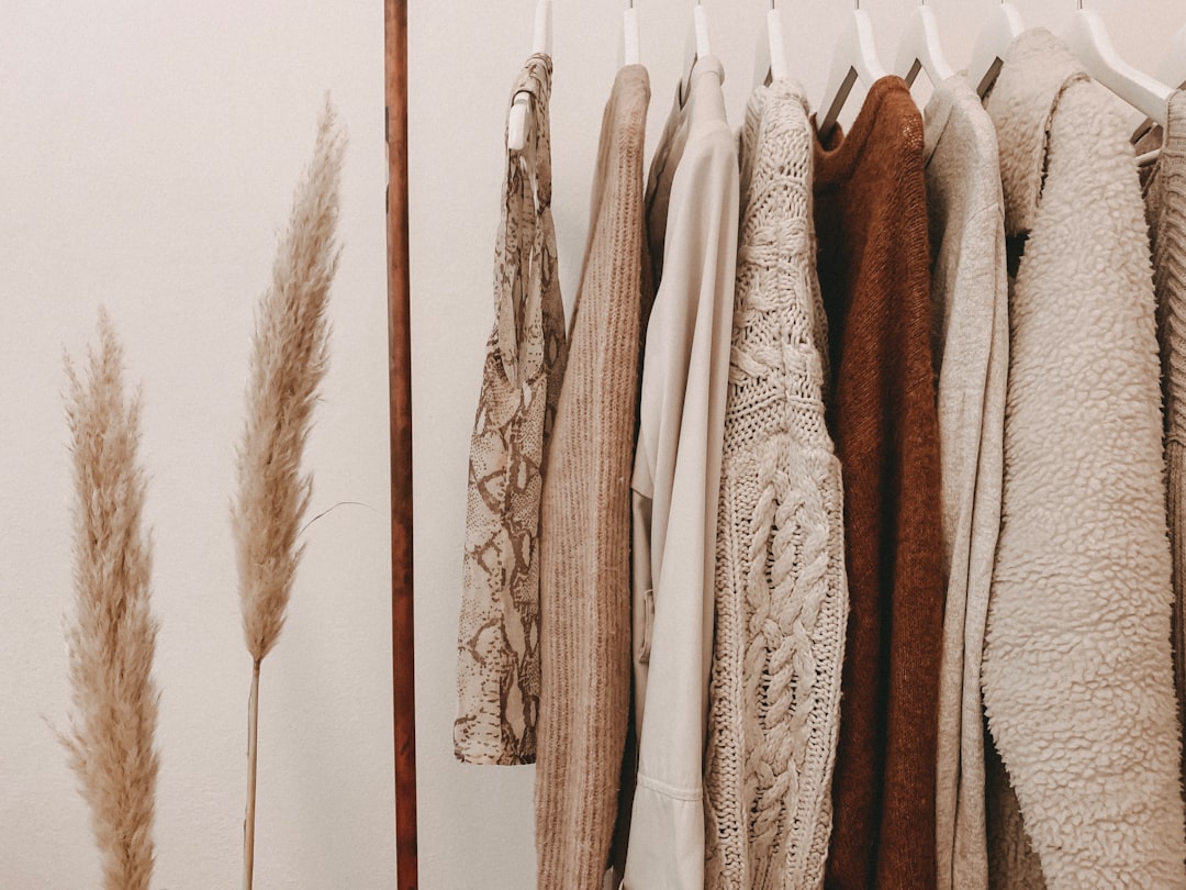 Clothing rack with assorted neutral-toned sweaters, cardigans, and tops hanging on white hangers, next to two pampas grass stalks against a light wall—an effortless nod to French-Girl Beauty.