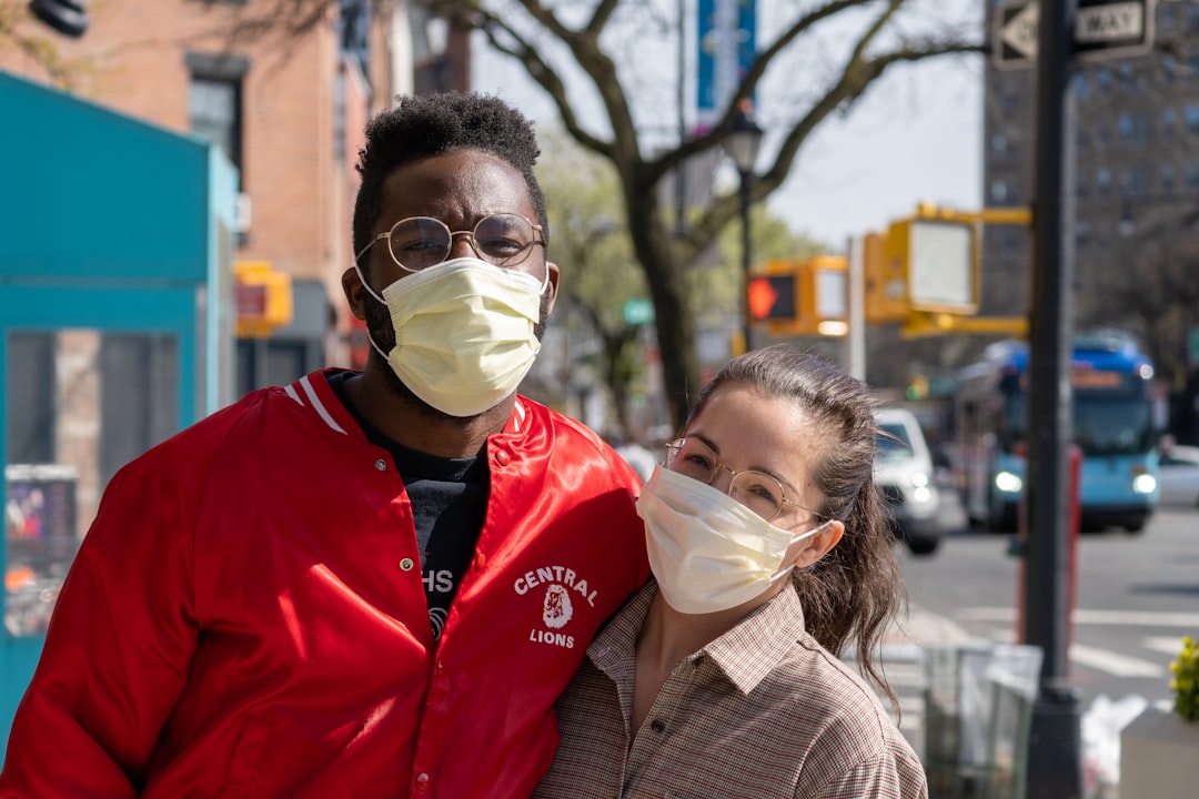 Two people wearing face masks stand close together on a city street during daytime, perhaps discussing Korean skincare rituals. One wears a red jacket, the other a plaid shirt. Urban background with trees and traffic signals.