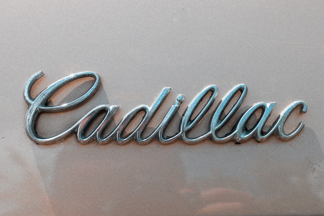 Silver Cadillac logo in cursive script showcased on a beige background, likely on the exterior of a car, giving it a jewelry-like elegance.