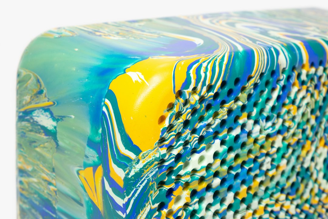 Close-up of a rectangular object with wavy, multicolored layers in blue, yellow, white, and green—this must-have piece features rounded edges and a row of small holes along one side, perfect for any fashionista’s limited edition jewelry collection.