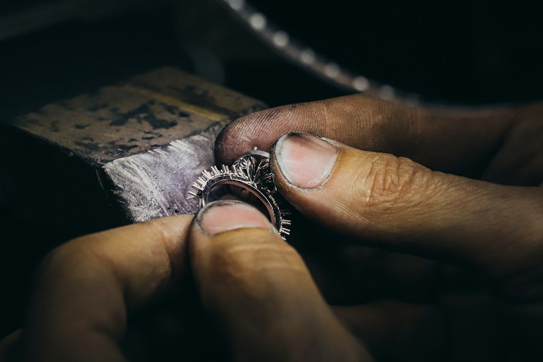 A close-up of hands working on fine jewelry, holding a ring and making detailed adjustments with visible tools and rough fingernails, capturing the craftsmanship behind each perfect piece.