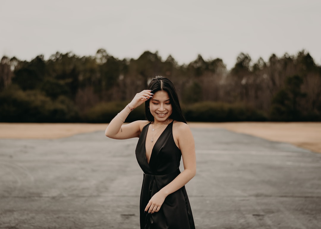 A woman in a black dress stands outdoors on a paved area, smiling and looking down, as she gets ready to unwind amid trees and a cloudy sky in the background.