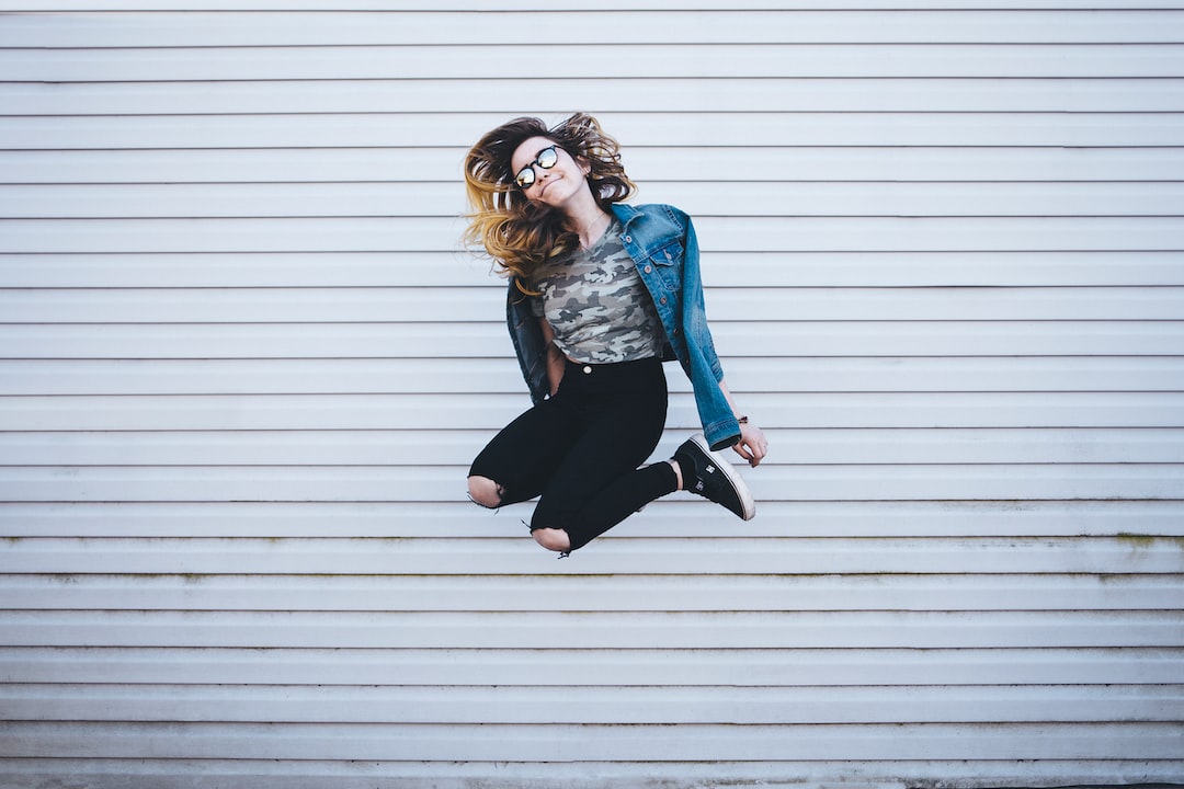 Young woman wearing glasses, a denim jacket, and ripped black jeans jumps in the air in front of a white, horizontal-striped wall, channeling punk influence with a nod to high fashion style.