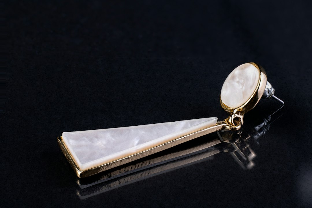 A single gold and white geometric drop earring with a triangular pendant and oval stud, displayed on a black surface, evokes the refined elegance of an exclusive ryokan Tokyo experience.