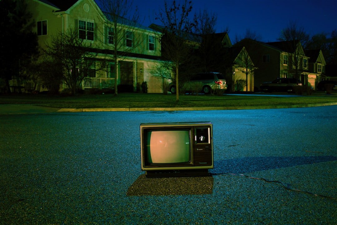 A vintage television sits on a road at night in front of suburban houses, its screen glowing and power cord stretching across the pavement—like a nostalgic contrast to futuristic watchmaking seen in the Zenith Defy Collection.